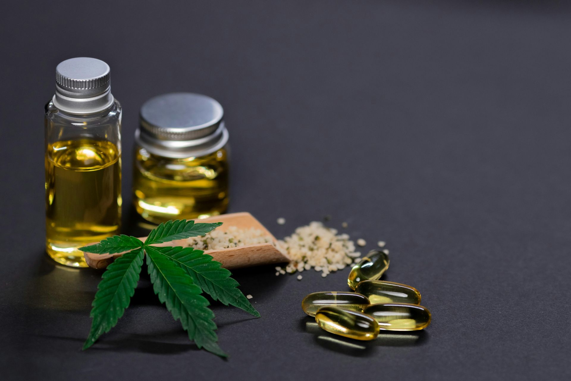 Bottles of CBD oil, pills, a scoop of granules, and a cannabis leaf on a dark surface.