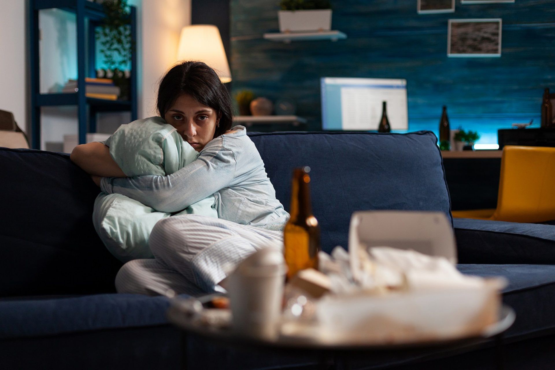 Woman on couch, hugging pillow, looks sad; table with tissues and bottle of beer in front.