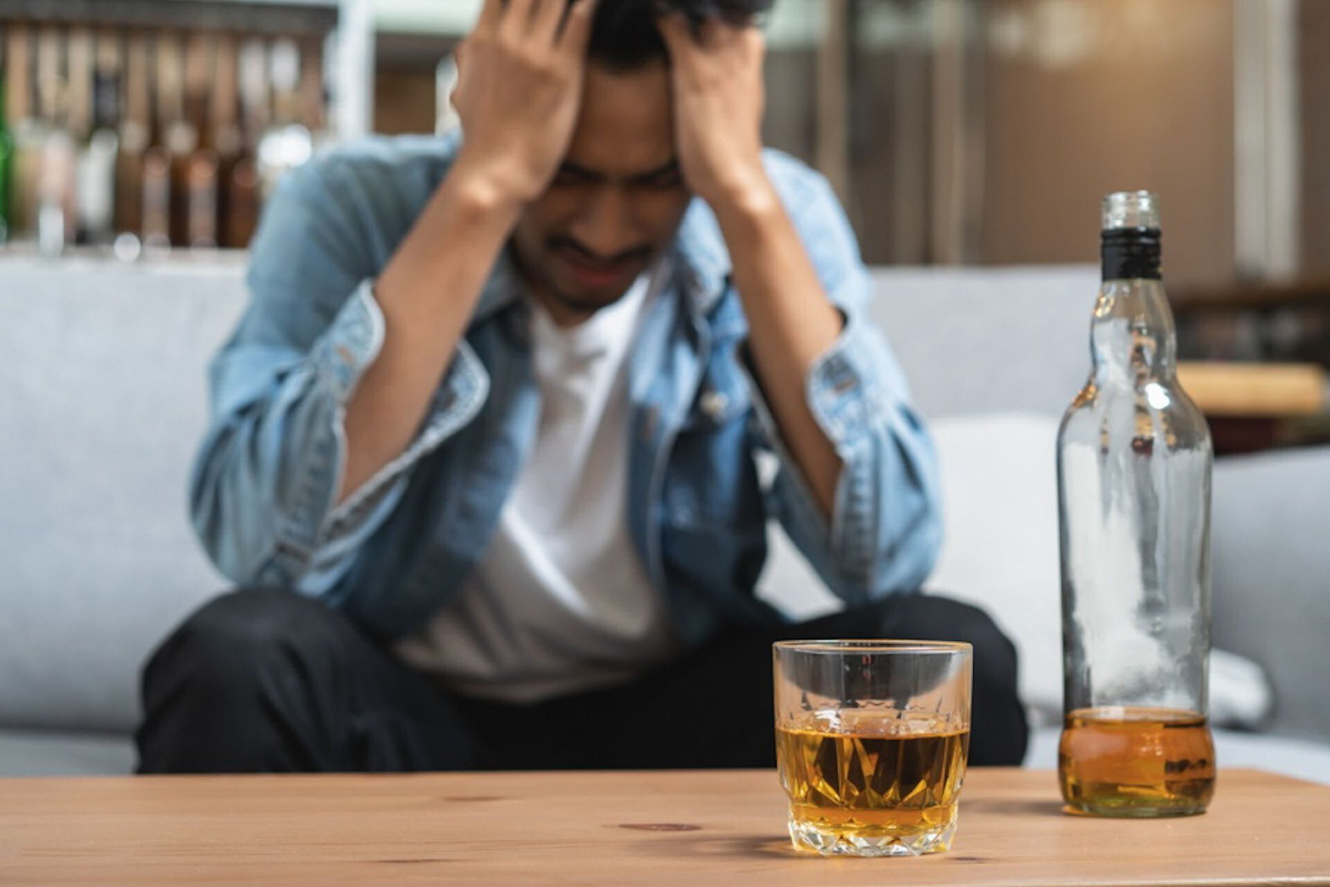 Man with head in hands, bottle and glass of alcohol on table.