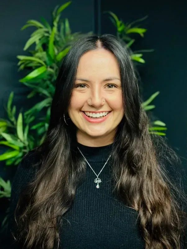 Woman with long brown hair smiles, wearing a black sweater, with a plant in the background.