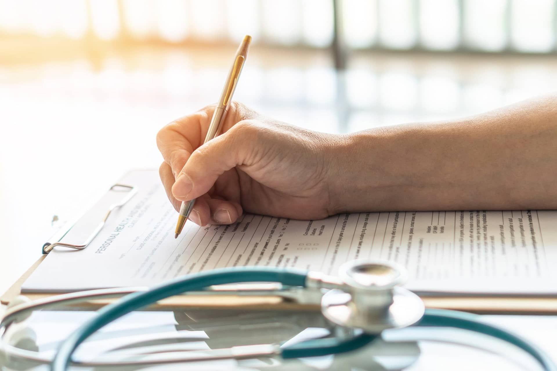 Hand writing on a clipboard with a stethoscope; sunlight shines on the scene.