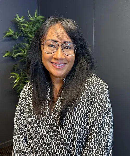 Woman with glasses smiling, wearing a black and white patterned shirt, seated in front of a plant and dark wall.