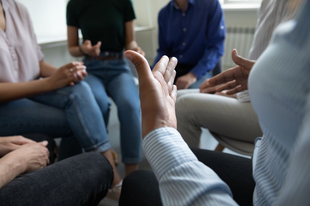 People in a circle, one person's hand raised, suggesting a discussion or therapy session.