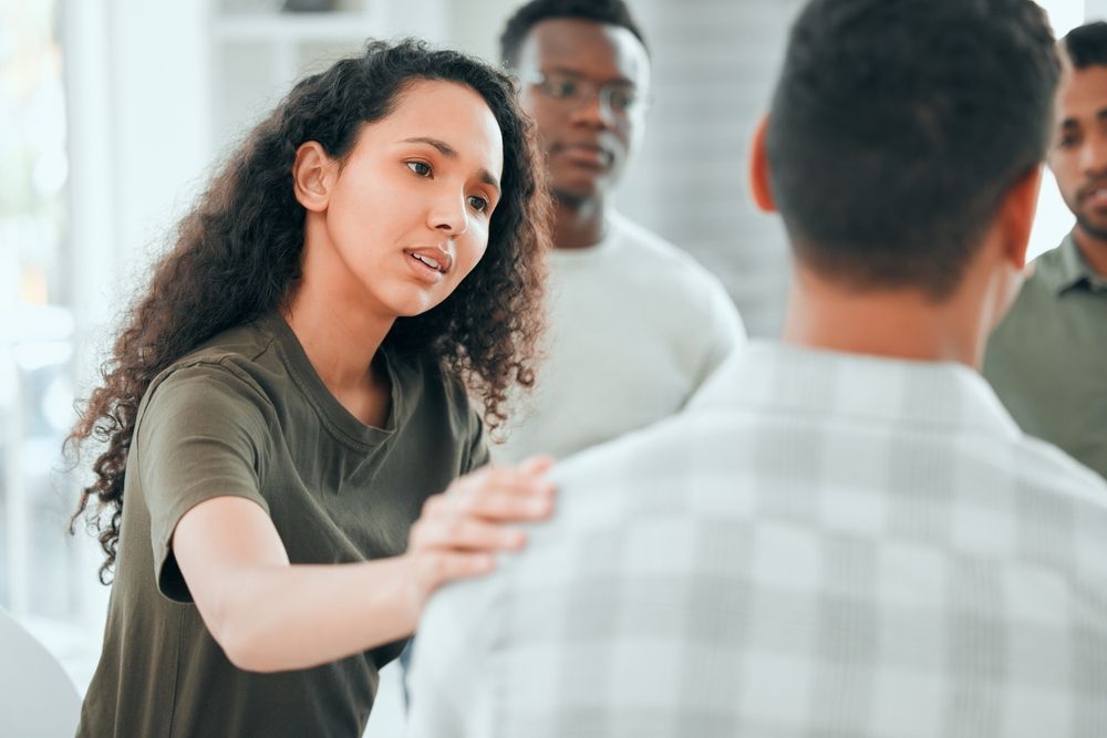 Woman comforts a man in a support group setting, hand on his shoulder, other members in the background.