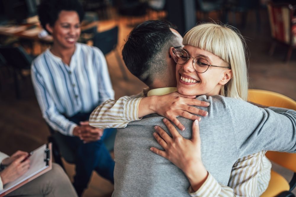 Couple hugging warmly in therapy session, smiling, with therapist in background.