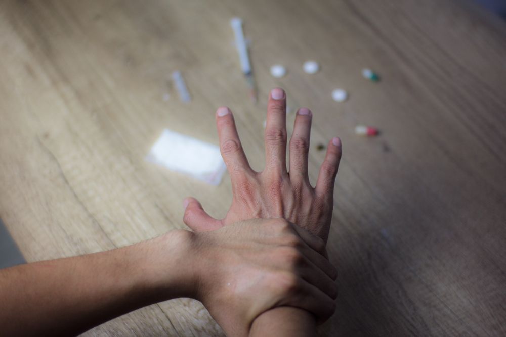 Two hands, one grasping the other's wrist. Pills, syringes, and a tissue are on a wooden surface.
