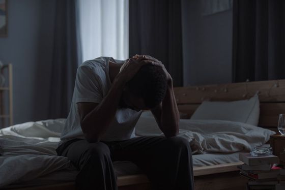 Man sits on bed, head in hands, looking distressed. Bedroom, low lighting.