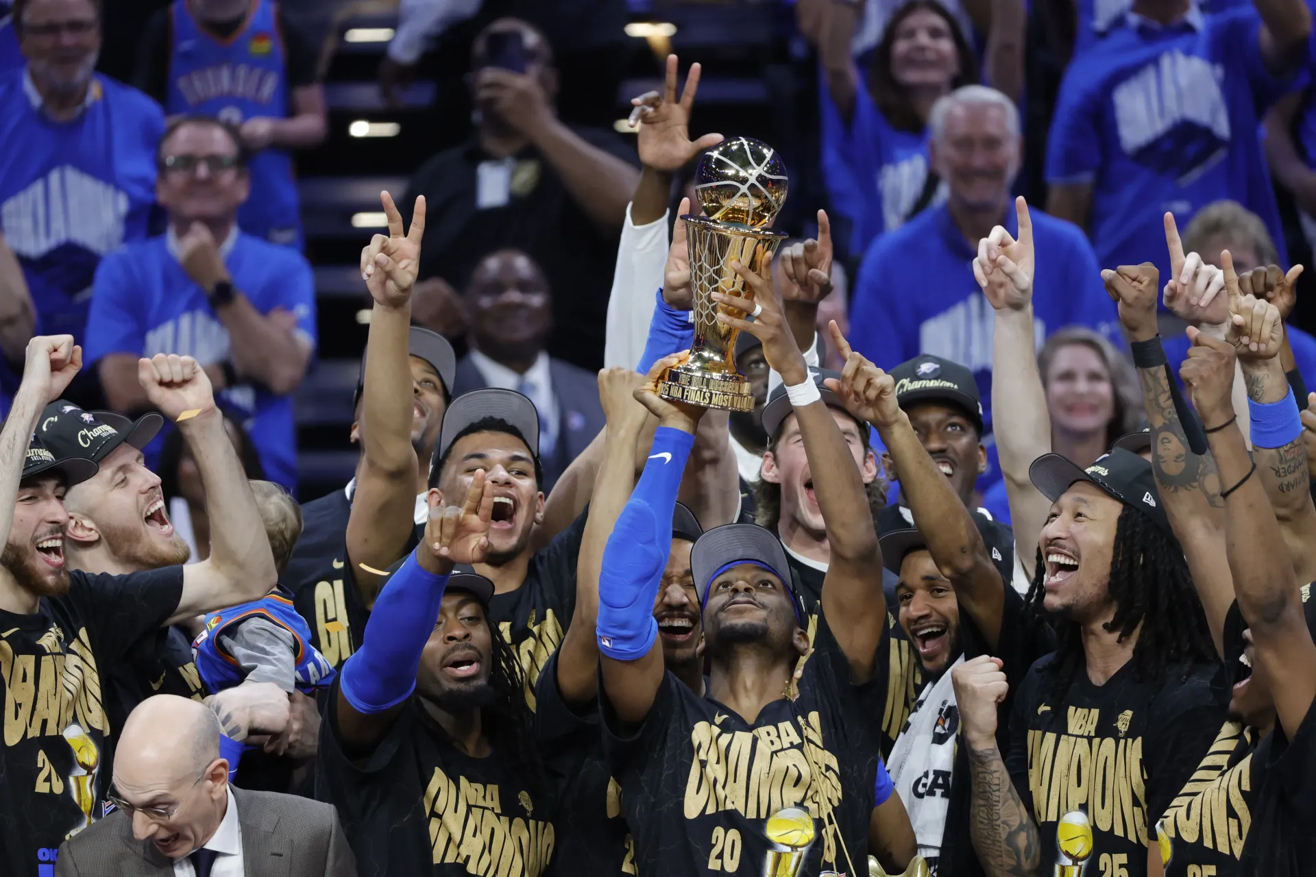 Oklahoma City Thunder players celebrate on the court, raising a gold trophy while wearing championship t-shirts.