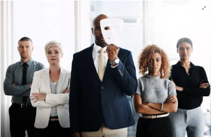 A man is holding a mask in front of his face while standing in front of a group of people.