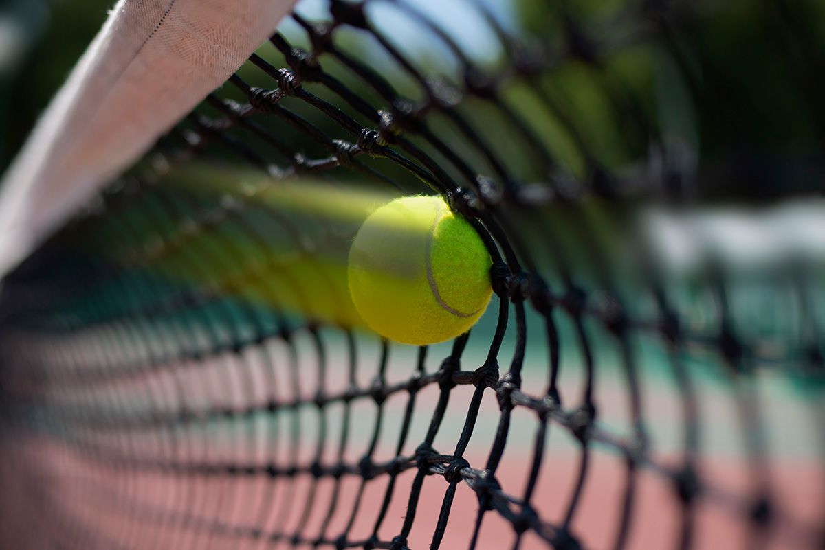 A bright yellow tennis ball strikes a black net on a tennis court, captured in motion with a blurred background.