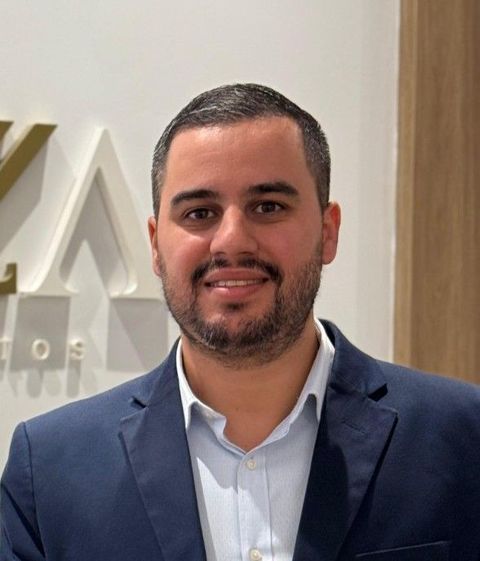 Homem de blazer azul-marinho e camisa azul-clara sorrindo em ambiente interno contra um fundo neutro.