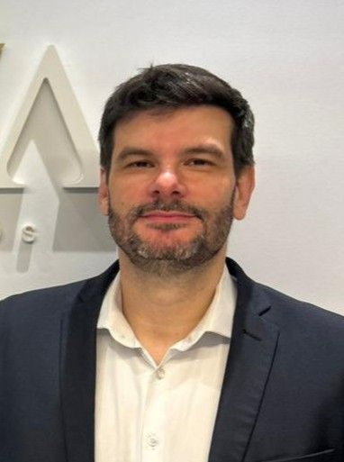 Homem de blazer escuro e camisa branca em pé dentro de casa contra uma parede clara com decoração geométrica.