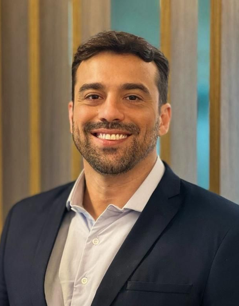 Homem sorridente de blazer escuro e camisa clara posando em ambiente interno contra um fundo neutro desfocado.