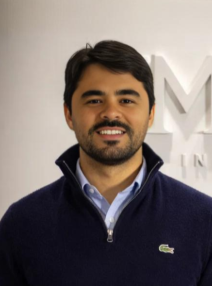 Pessoa sorridente vestindo um suéter azul-marinho, em pé dentro de casa contra uma parede clara.