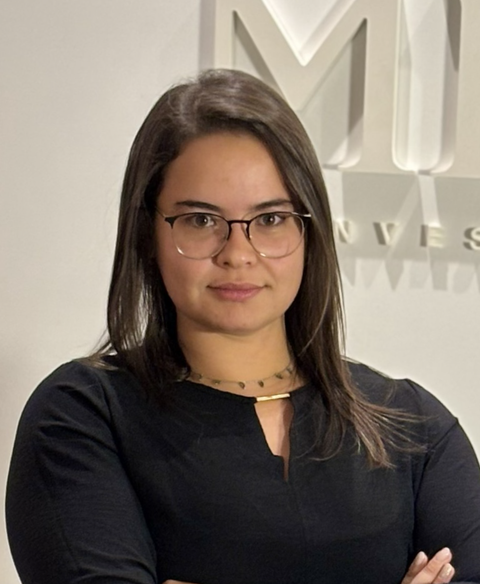 Mulher de óculos em pé, com os braços cruzados, em frente a uma parede com um grande logotipo.