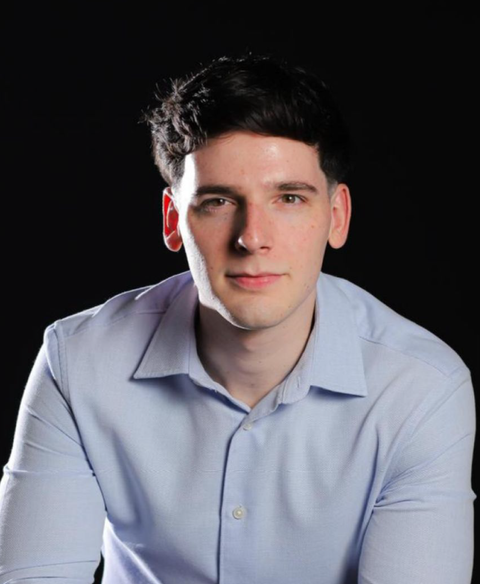 Retrato de uma pessoa com camisa azul clara contra um fundo preto, olhando para a câmera.