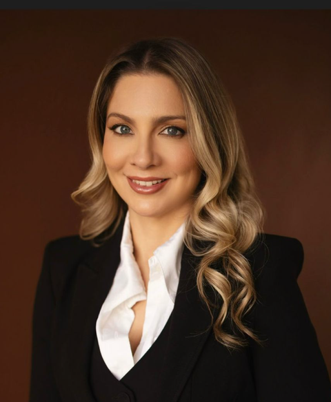 Retrato de uma pessoa sorridente vestindo um blazer preto e uma blusa branca contra um fundo marrom.