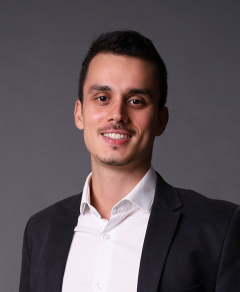 Retrato de uma pessoa sorridente vestindo um blazer escuro e camisa branca contra um fundo cinza.