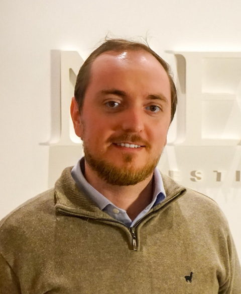 Homem de suéter bege parado dentro de casa diante de uma parede branca.