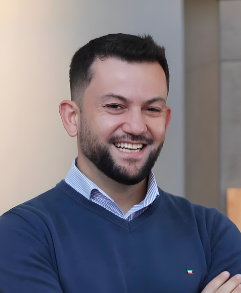 Homem sorridente de suéter azul, em pé dentro de casa com os braços cruzados.