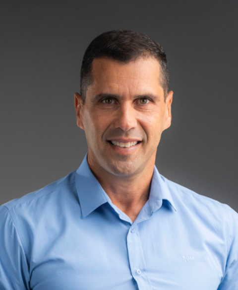 Retrato de um homem sorridente com uma camisa azul-clara de botões contra um fundo cinza.