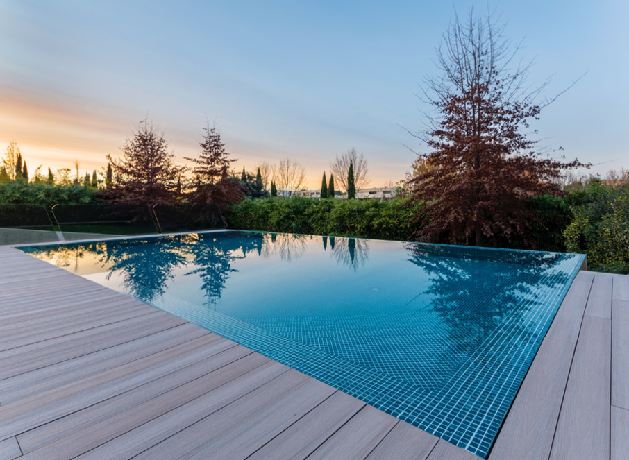 An infinity pool reflects a sunset sky; light wooden deck on the left.