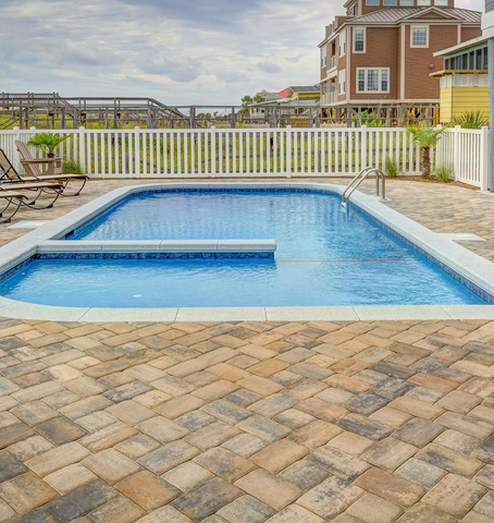 Rectangular in-ground pool surrounded by brick patio. White fence and houses in the background under cloudy sky.