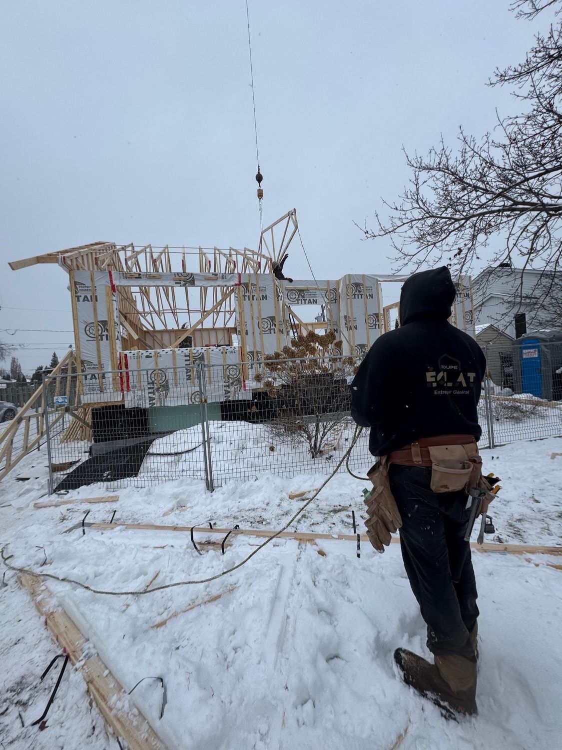 Un ouvrier du bâtiment regarde la charpente d'une maison soulevée par une grue dans un décor enneigé.