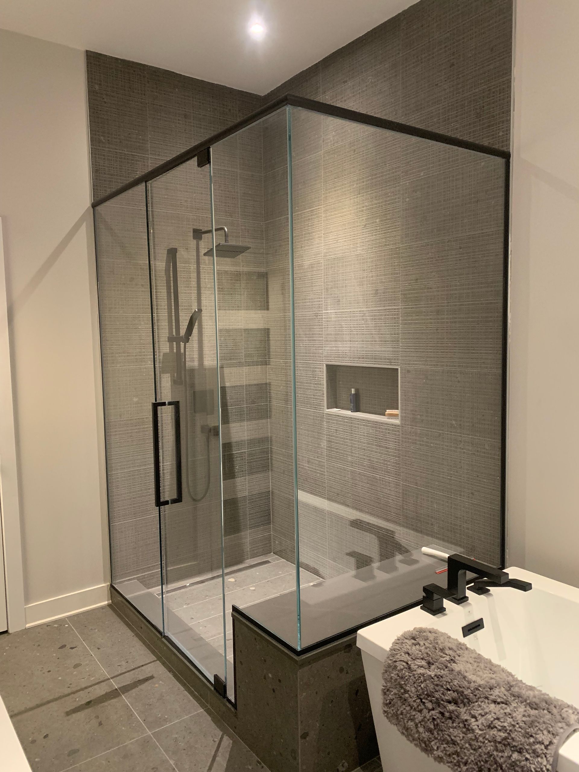 Cabine de douche moderne avec cadre noir et carrelage gris texturé. Une baignoire est visible à droite.