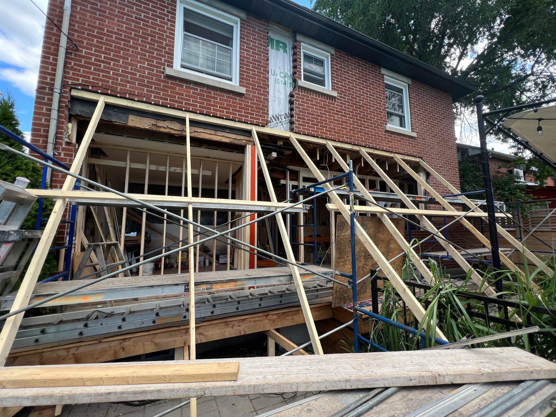 Exterior of a brick house under construction; wooden framing covers the former porch area, visible scaffolding.