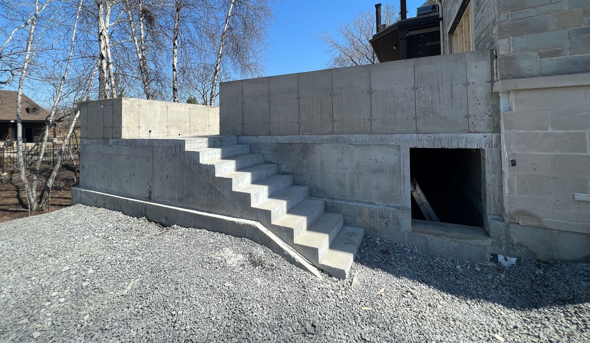 Escalier en béton menant à un mur en béton à côté d'une porte; sol en gravier, arbres nus.