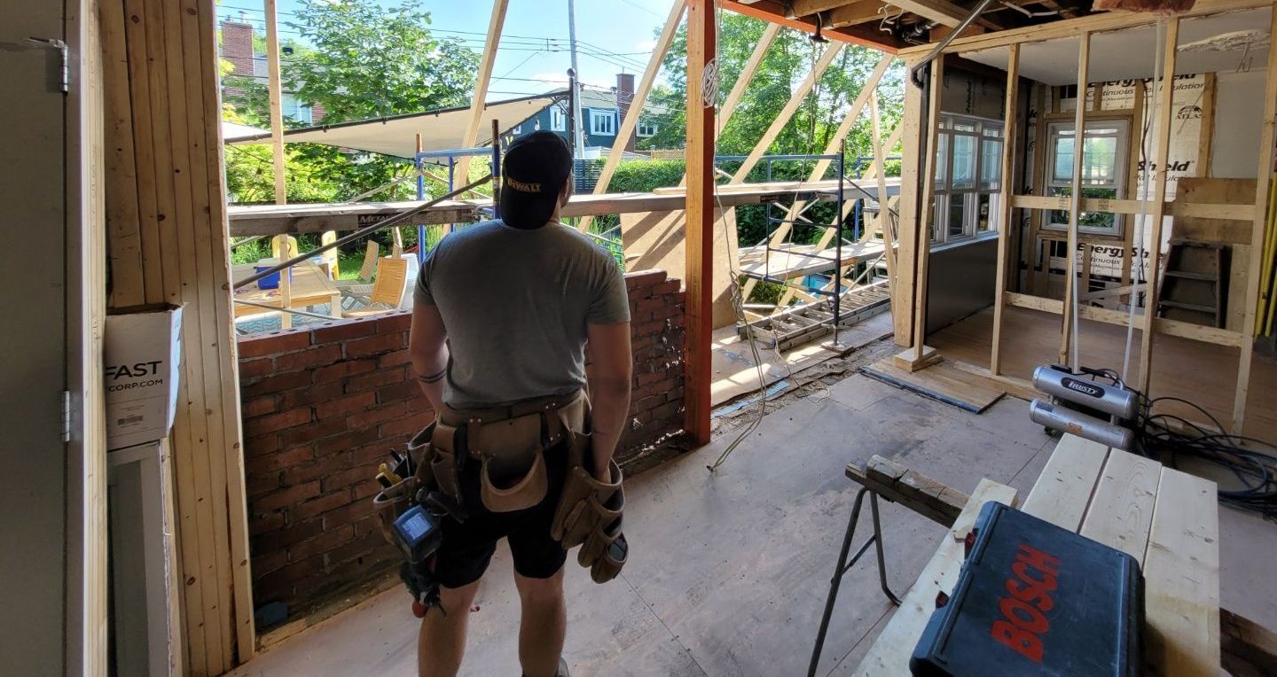 Un homme, ceinture à outils à la main, regarde depuis un bâtiment en construction. On aperçoit des échafaudages.