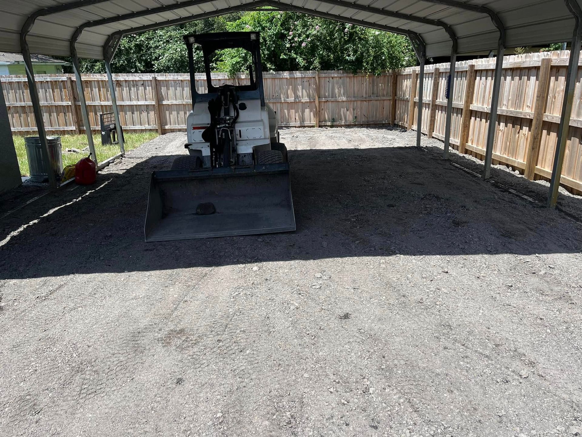 Bobcat tractor parked on gravel under a carport, fenced backyard setting.