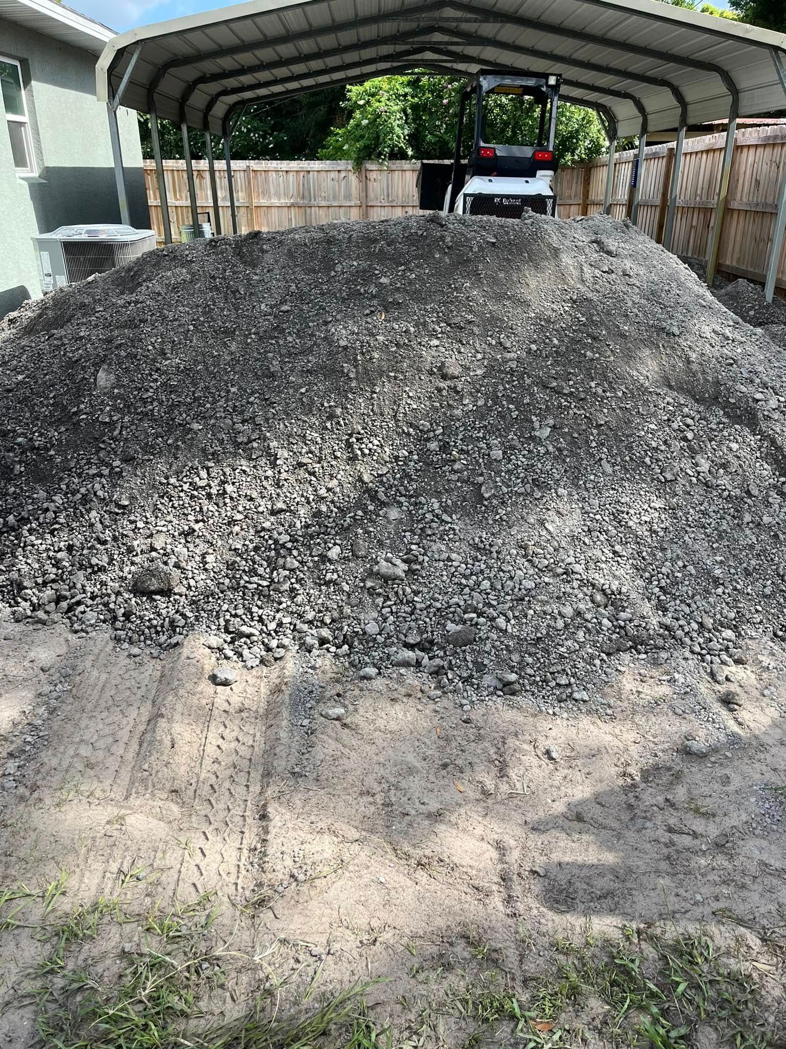 Large pile of dark gravel under a metal carport, with a small tractor visible in the background.