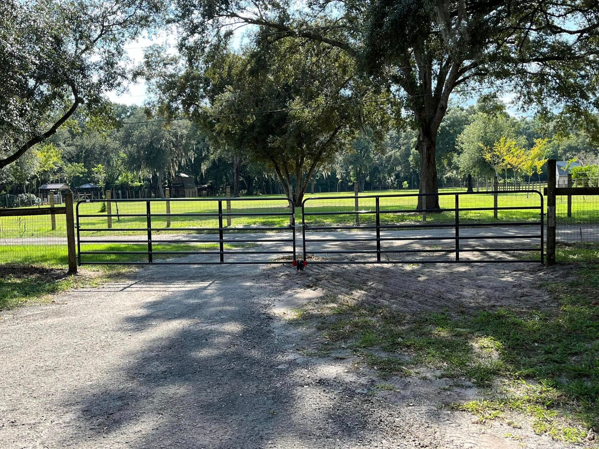 A gravel driveway leads to a closed metal gate in a rural setting with trees and green grass.