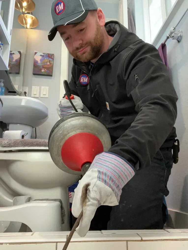 Plumber using a drain snake in a bathroom to unclog a drain, wearing gloves, cap and dark jacket.