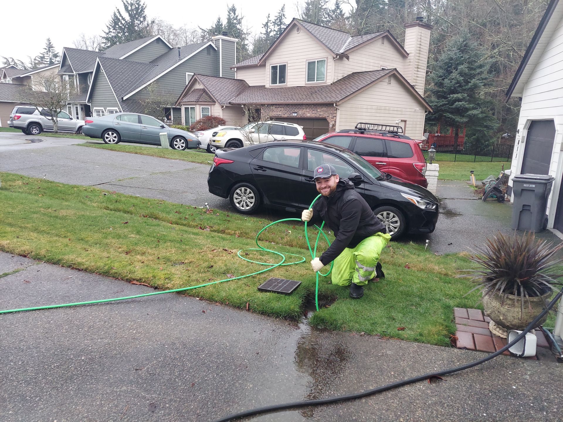 Person in neon green working on a drain in a residential driveway, holding a hose. Cars and houses are in the background.