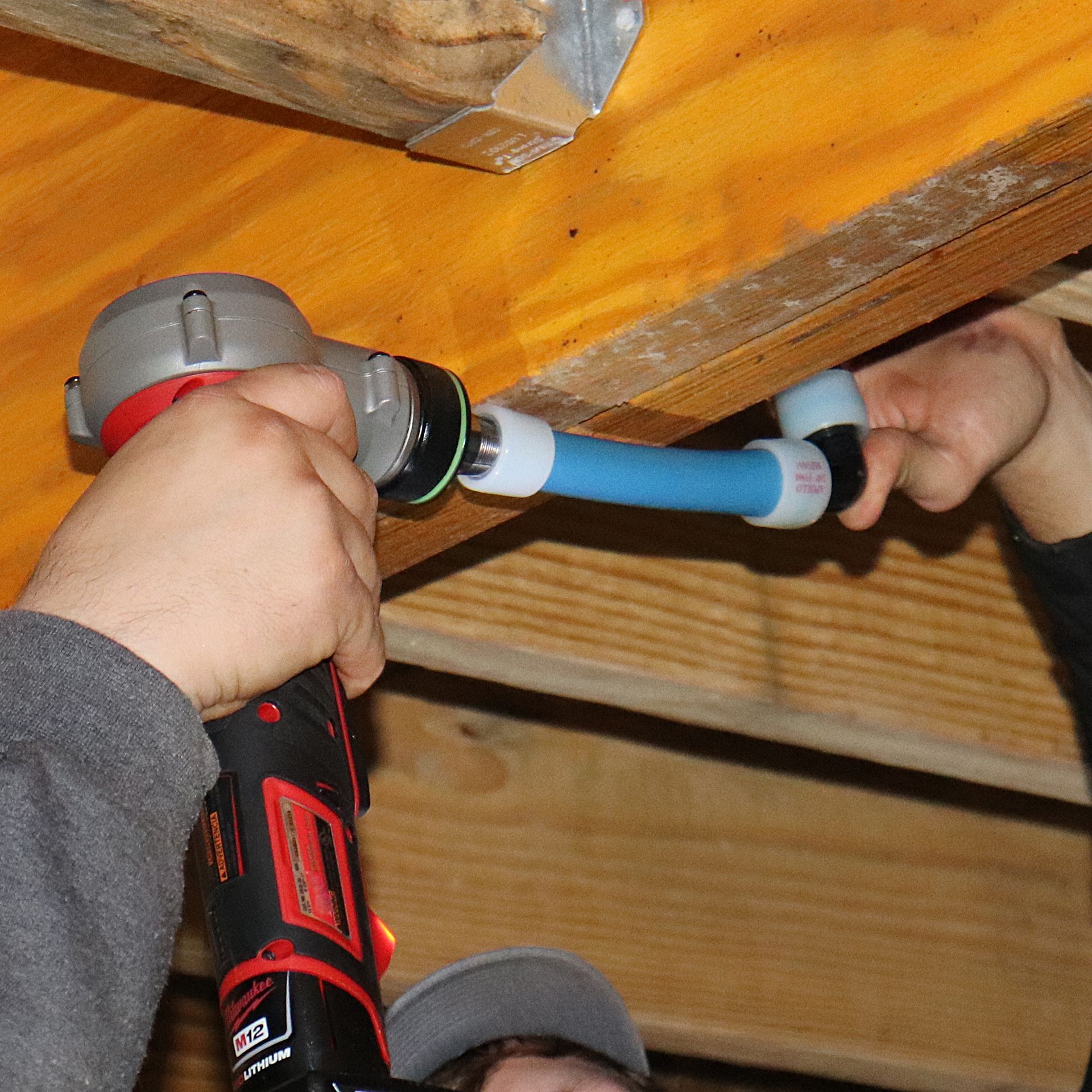 Person using a drill to connect blue PEX pipe and fittings in a wooden structure.