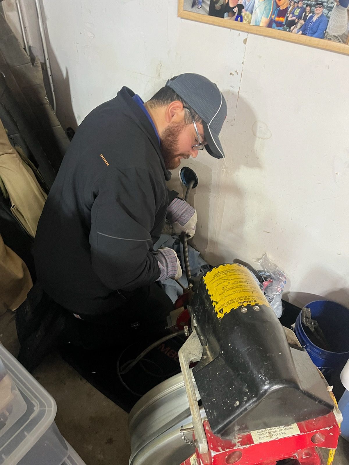 Man working on a machine in a garage. Wearing a cap and holding a tool, near a barrel.