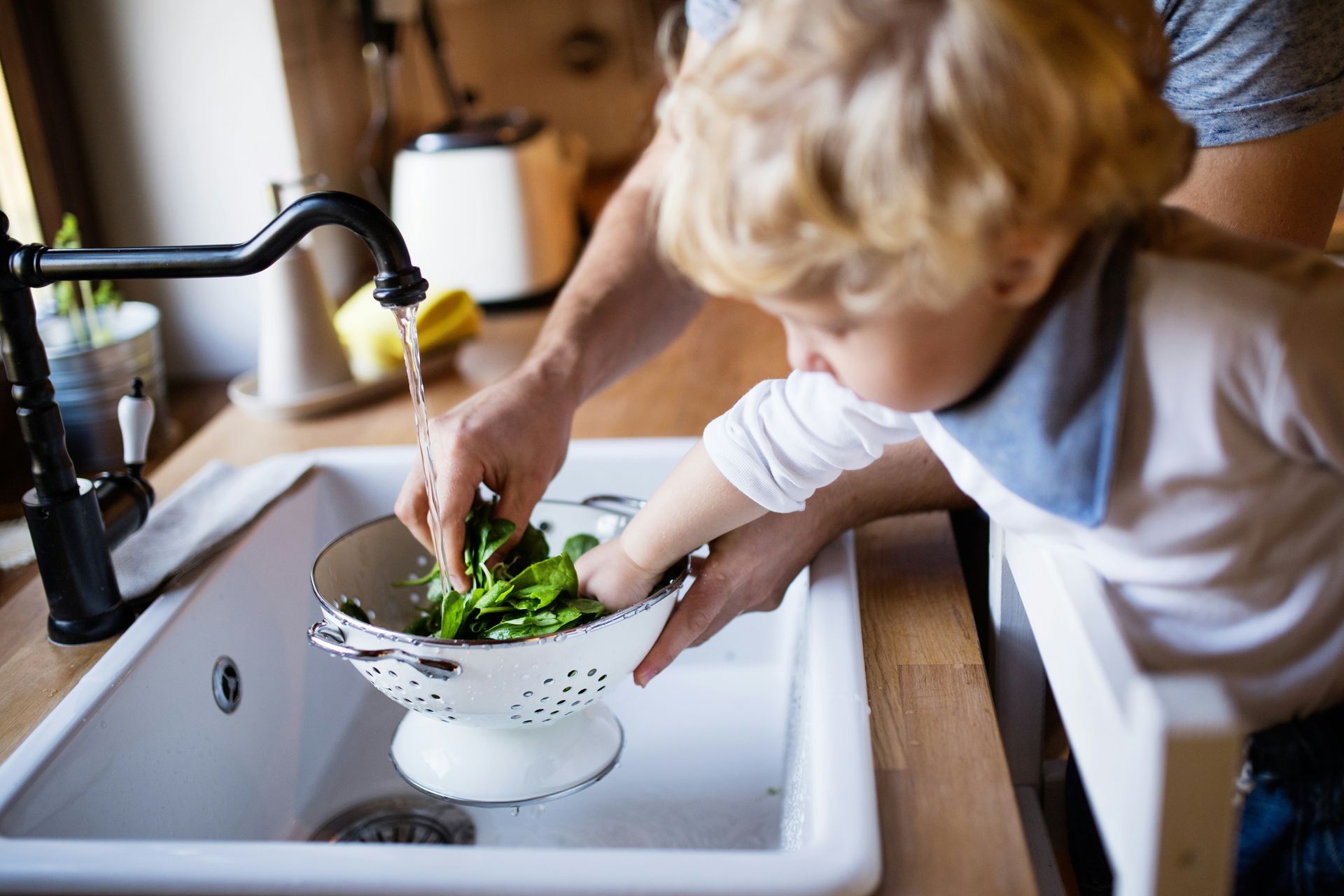 Child helping adult wash spinach in a kitchen sink.