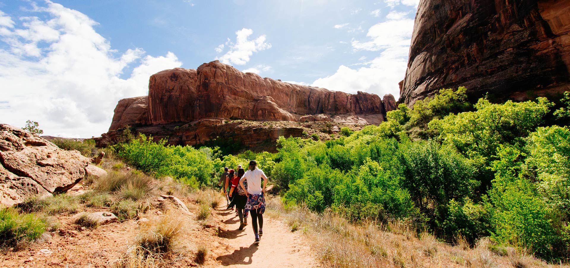 People hike on a trail under a blue sky, surrounded by red rock formations and green bushes.