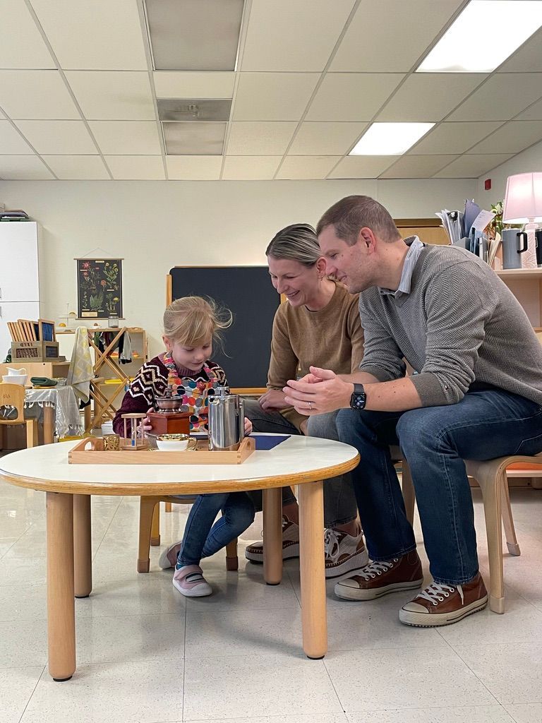 Parents smiling watching their daughter complete a Montessori lesson.