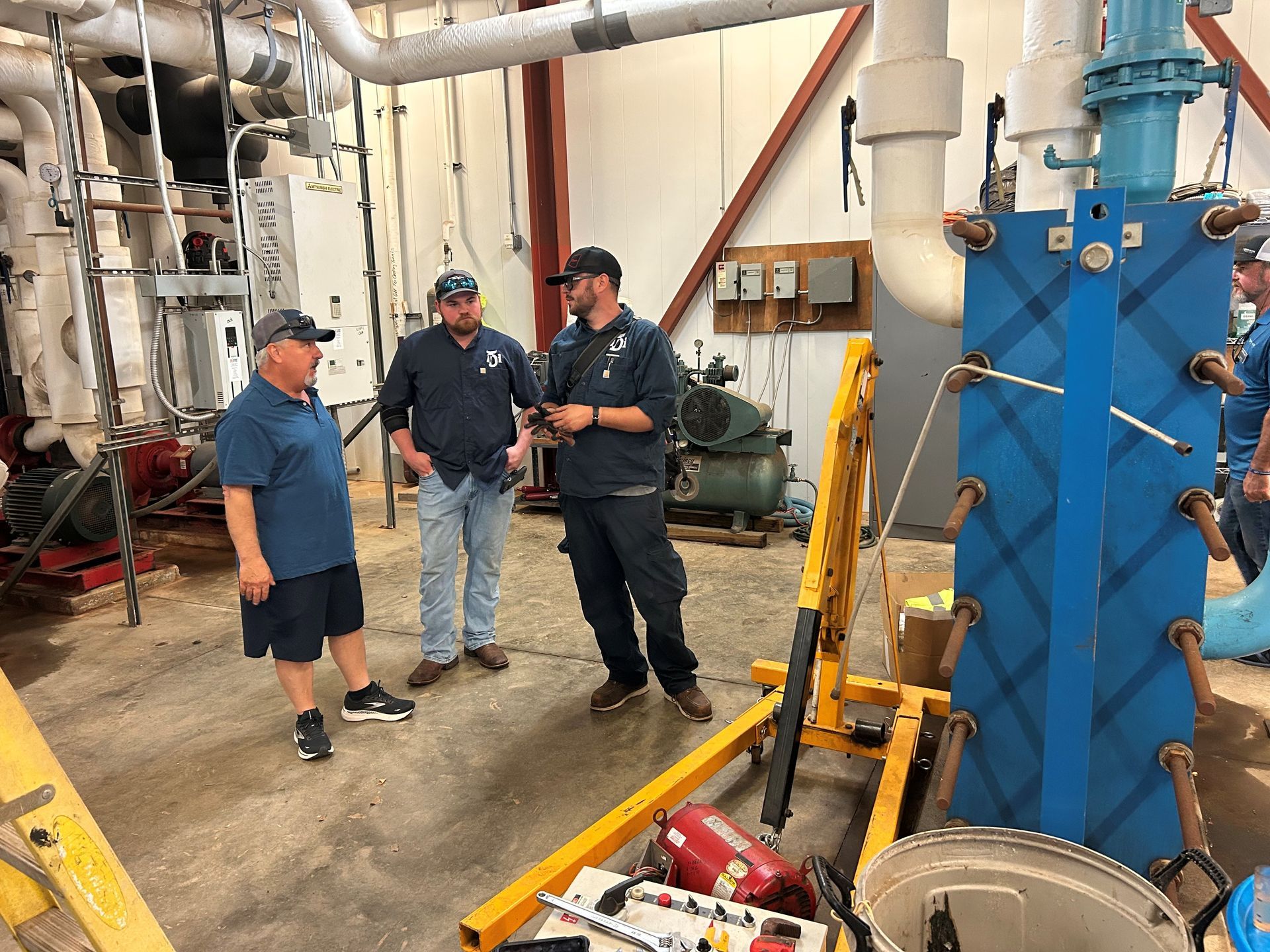 Three men discuss near blue machinery and pipes in a utility room.