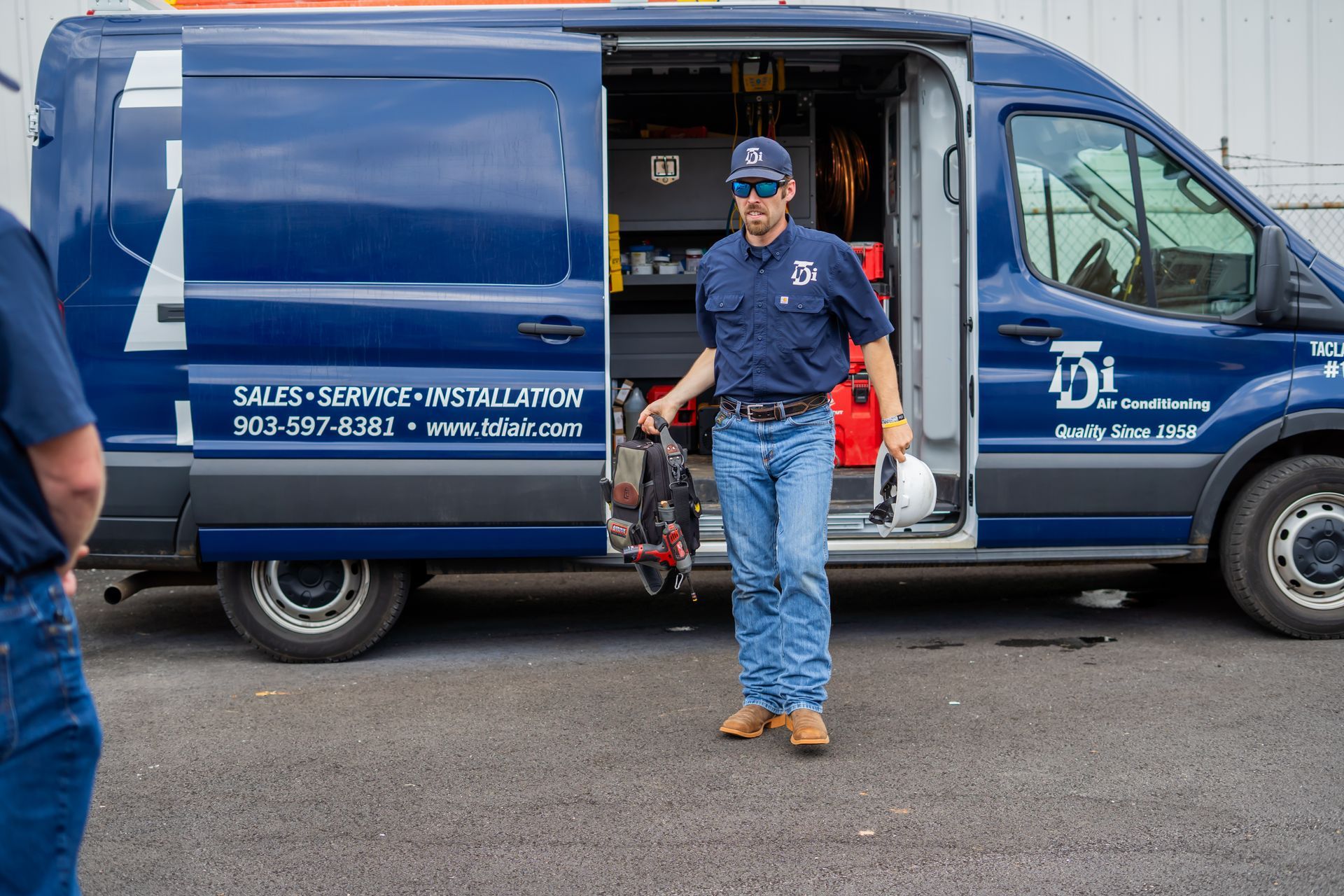 Man in blue work clothes holding equipment by a dark blue van, parked outside.