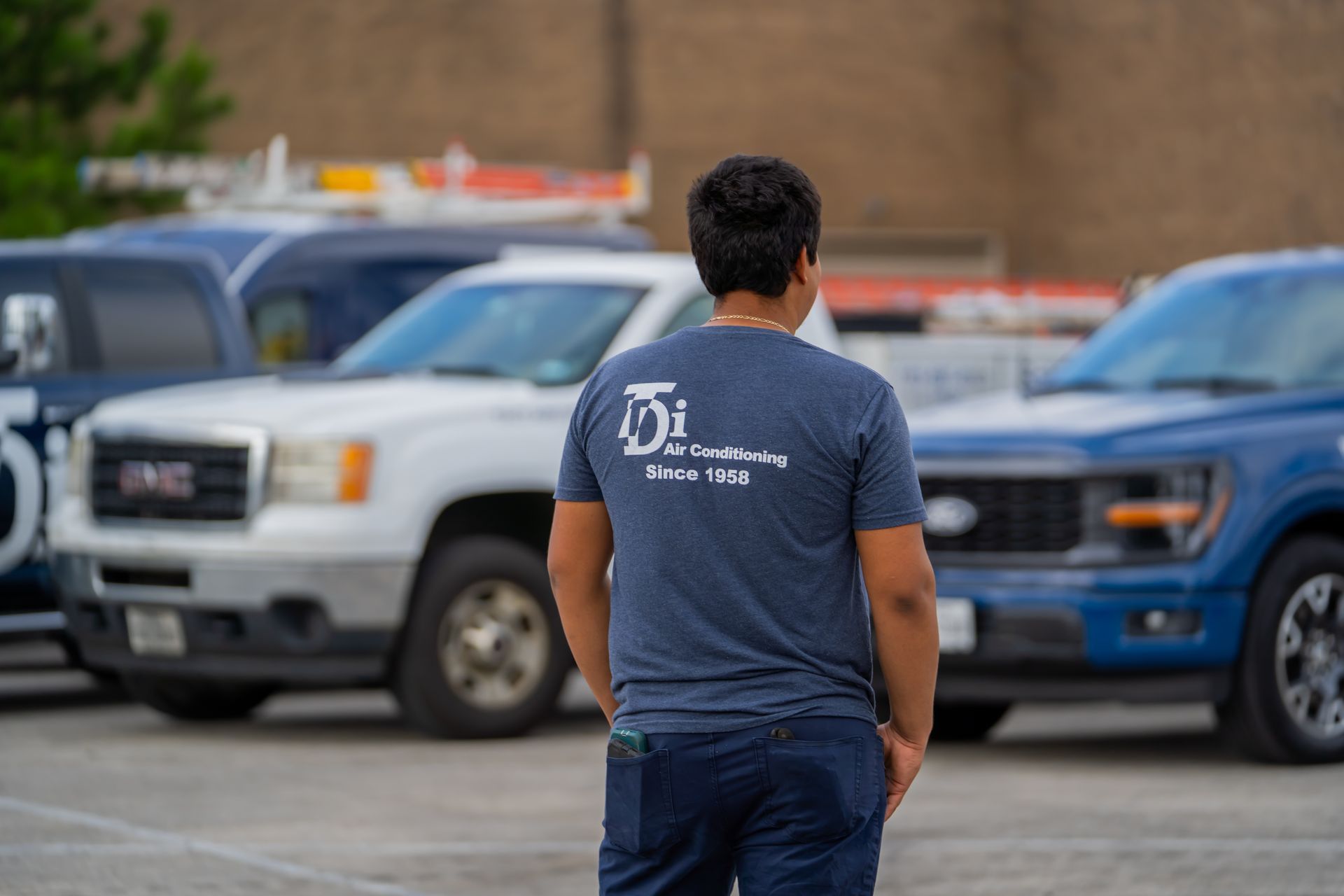 Man in blue shirt with company logo stands in front of service trucks in a parking lot.