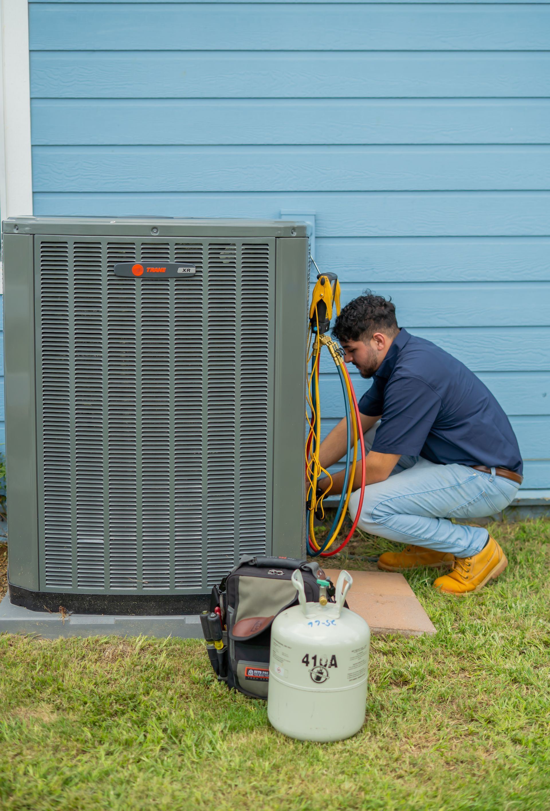 HVAC technician works on an air conditioner unit outside a blue house. Includes gauges, tools, and refrigerant tank.