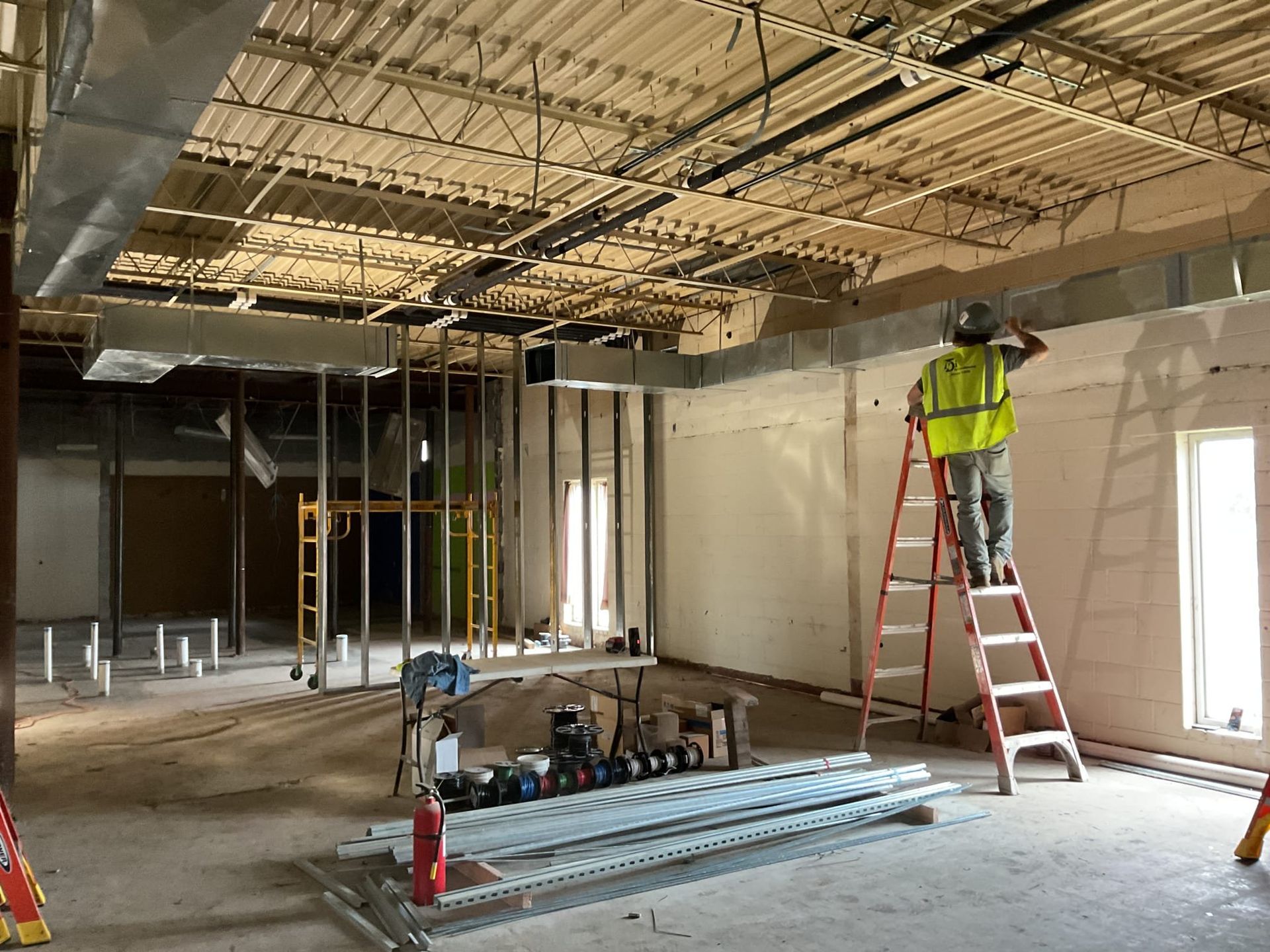 Construction worker on a ladder inside a building, wearing a safety vest, working on the ceiling.