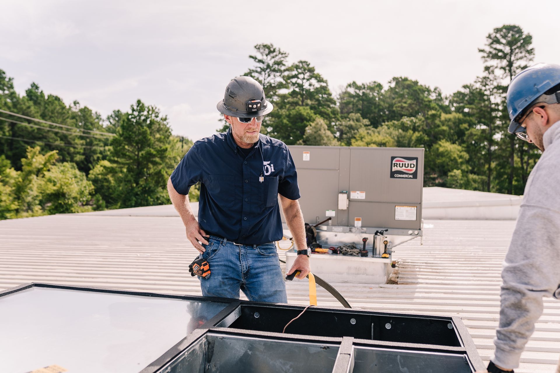 Two HVAC technicians on a rooftop inspect equipment; one in a hard hat, another in a blue cap.