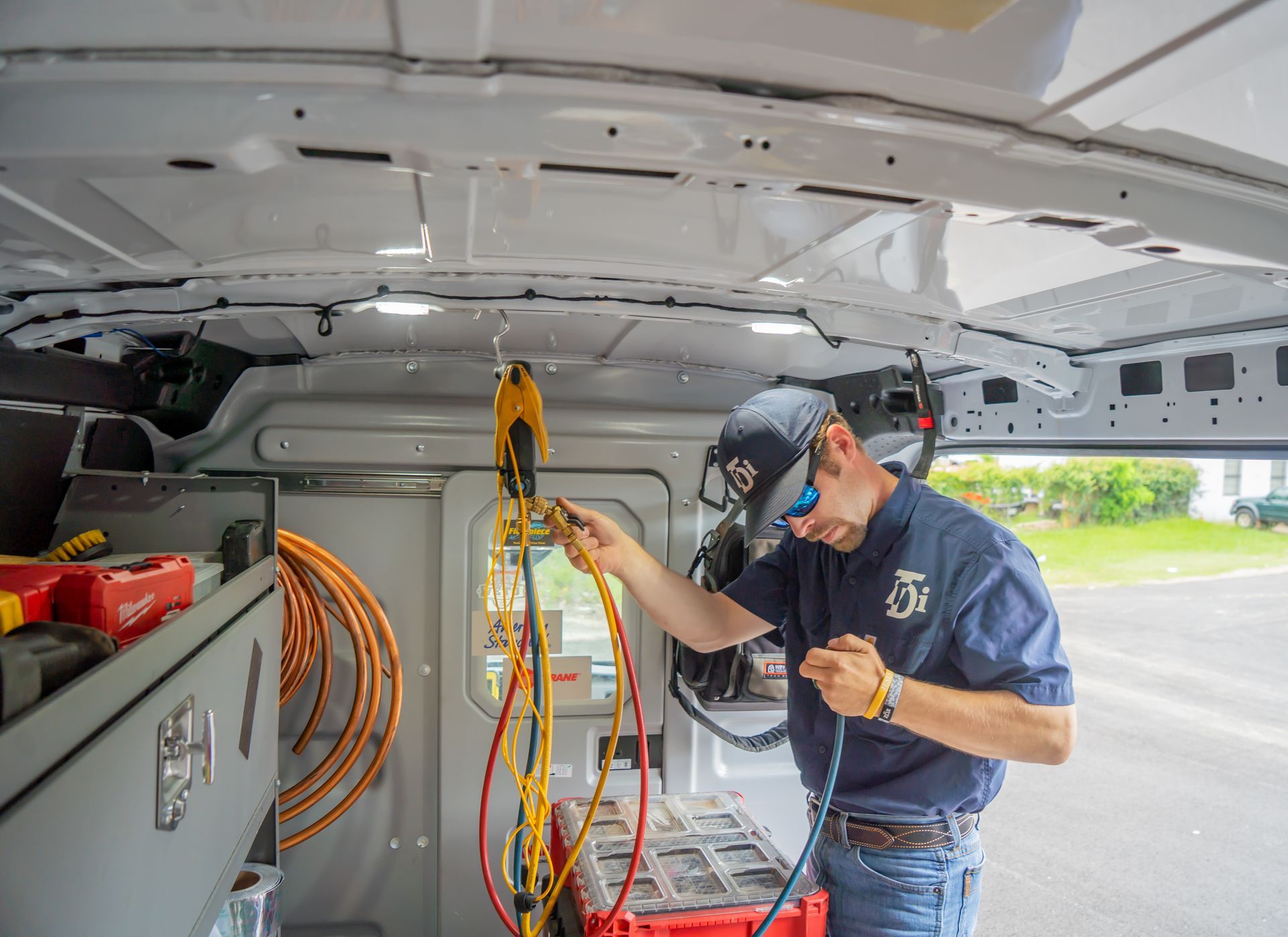 Man working inside a van, holding wires. He wears a blue shirt and cap. The van has organized tools.