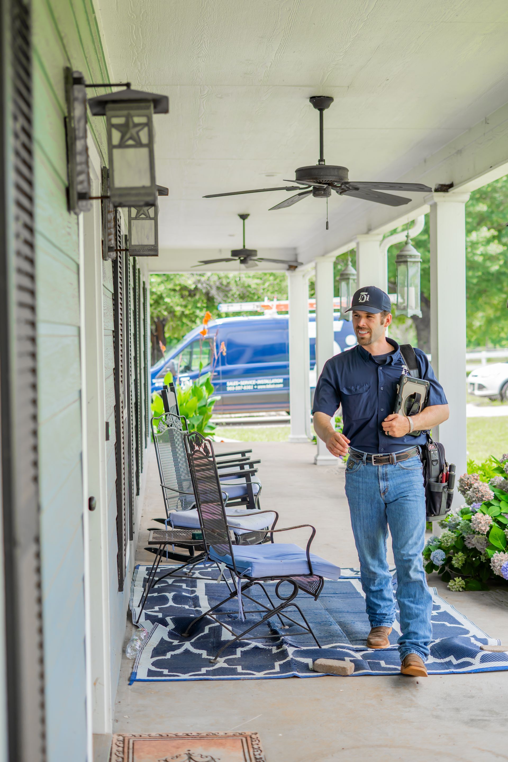 An electrician smiles on a porch, holding a device. A blue van is parked in the background.